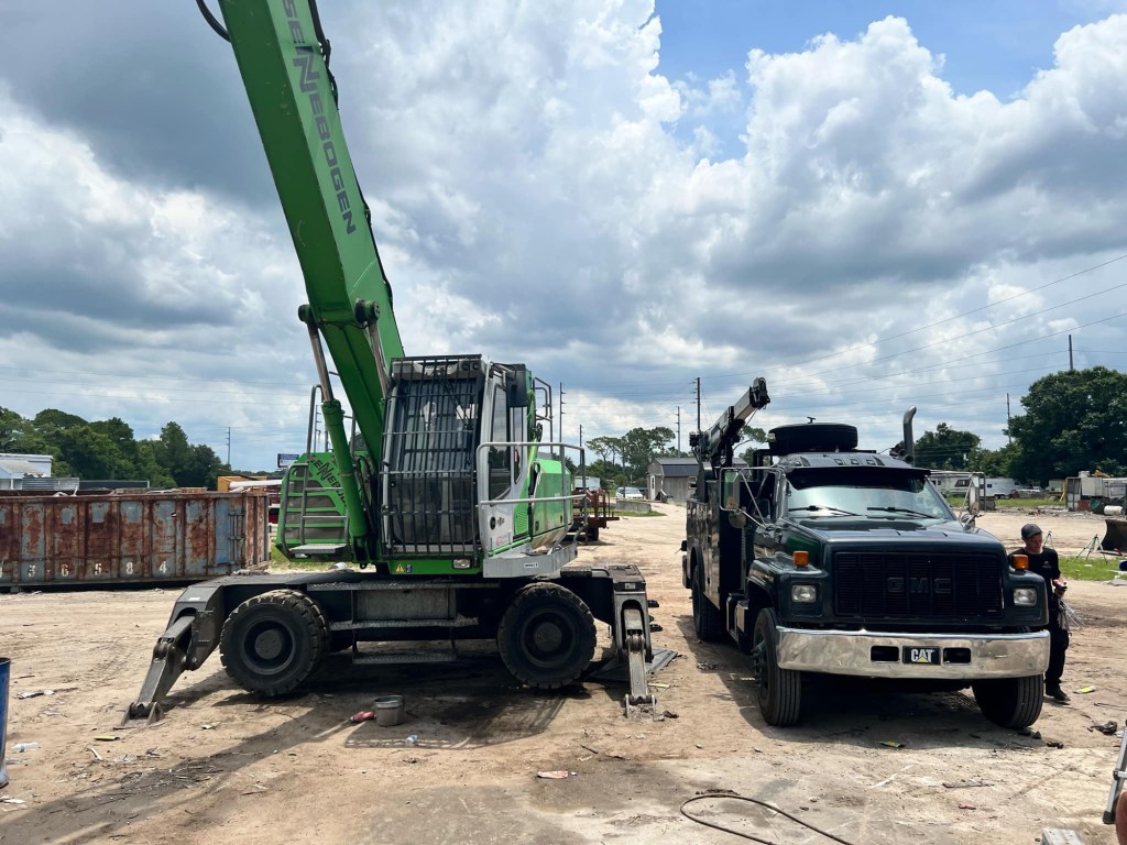 A green SENNEBOGEN machine is parked next to a dark-colored GMC truck in a heavy equipment yard, with scattered debris and containers in the background under a cloudy sky.